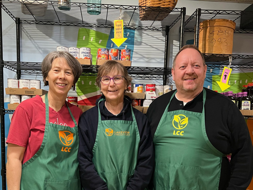 Three middle-aged people wearing aprons, pose smiling in front of shelves of grocery items.