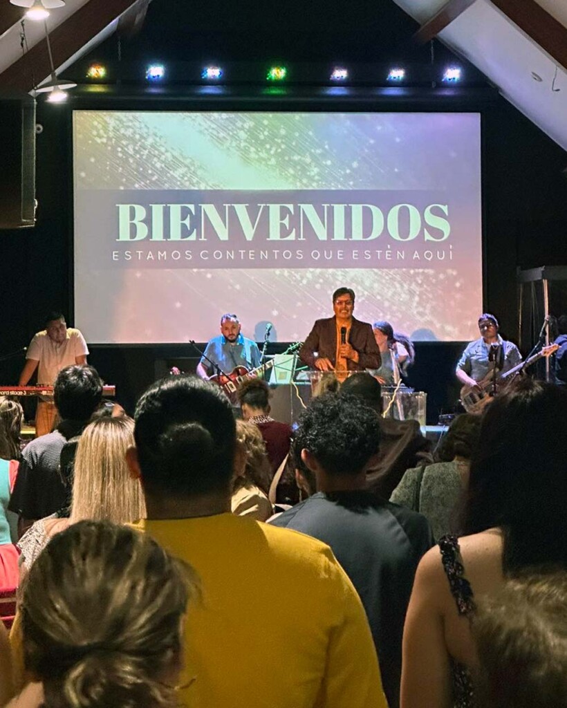 Pastor Filiberto, wearing a suit and holding a microphone, addresses a congregation. Behind him, is a large screen saying "Bienvenidos!", and several musicians wait with their instruments. The room is spacious, with multicolored lighting overhead.